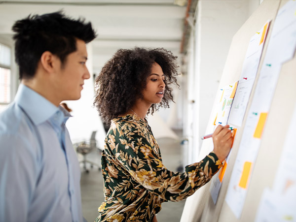 two people at a bulletin board with papers and sticky notes in an office setting