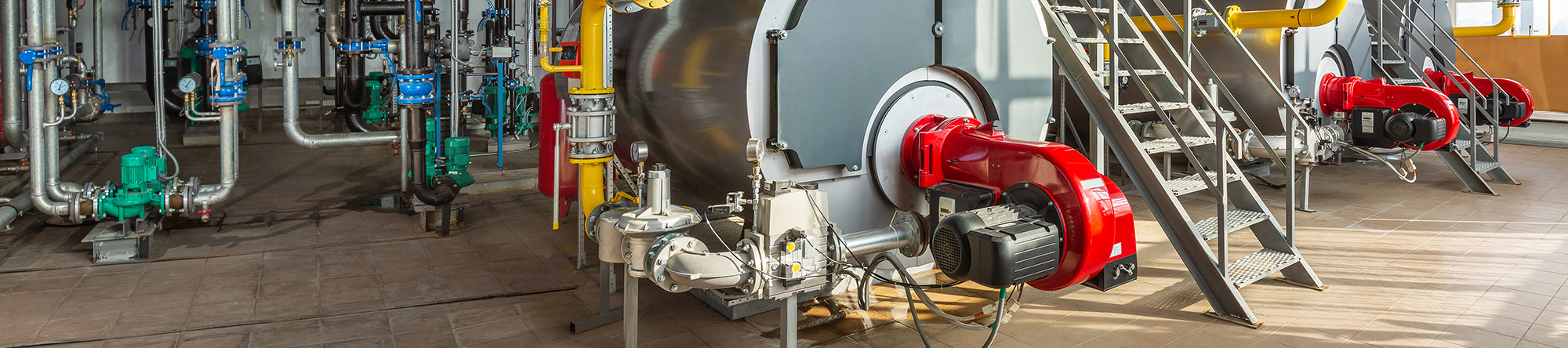 interior of an industrial facility featuring large machinery, interconnected pipes, red and green valves, yellow safety railings, metal stairs, and a grey cylindrical tank